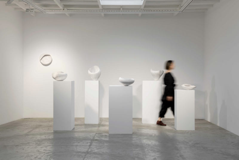 A gallery installation showing a series of white ceramic sculptures by Yasha Butler, displayed on tall white pedestals. The organic, curved forms rest on and float above the plinths against pristine white walls. A blurred figure in black clothing walks through the space, creating a sense of scale and movement. The minimalist presentation in the concrete-floored gallery space emphasizes the pure, sculptural forms of the artworks.